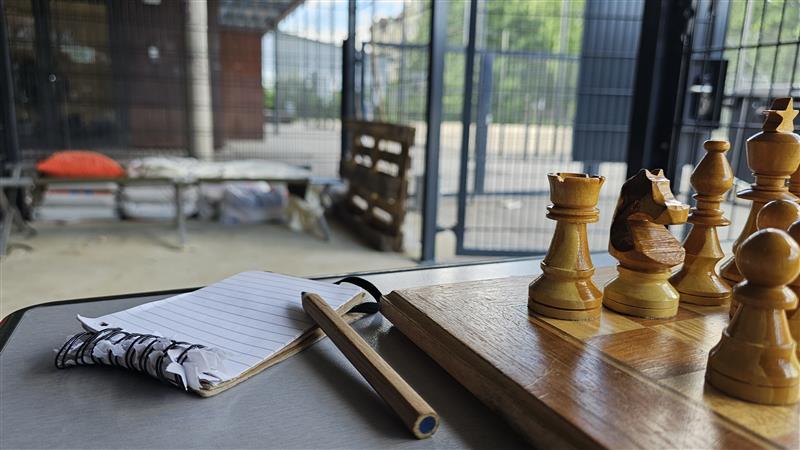Im Vordergrund ein Tisch. Darauf zu sehen eine Ecke eines Schachbretts mit aufgestellten Figuren. Daneben ein Notitzblock und ein Buntstifft. Im Hintergrund Gitterwände.