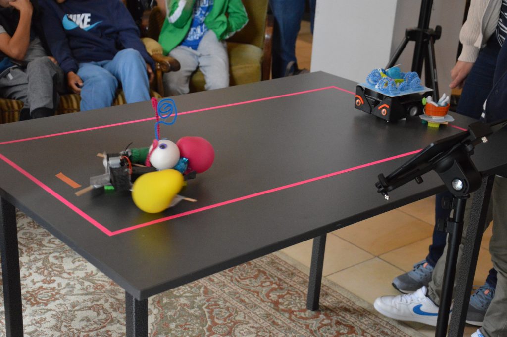 Auf einem schwarzen Tisch wurde mit rotem Klebeband ein rechteck abgeklebt. Darauf stehen zwei aus diversen Schrottteilen gebaute Roboter - bereit für den Wettkampf.