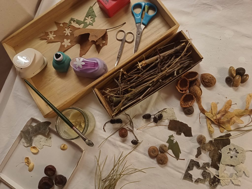 Oberfläche mit weißer Tischdecke. Darauf verschiedene Naturmaterialien, wie kleine Äste, Blätter, Kastanien, Eicheln, Nussschalen. Aus einigen Blättern wurden Muster ausgestanzt. Außerdem zwei scheren, einige Stanzer und ein Glaß auf dem ein Pinsel liegt.