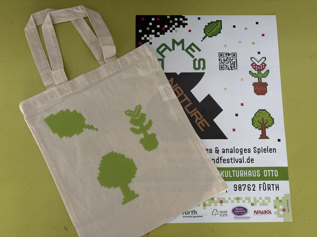 Jute-Tasche mit darauf geplotteten Festival-Symbolen liegt mit einem Festival-Plakat auf einer grünen Oberfläche.
